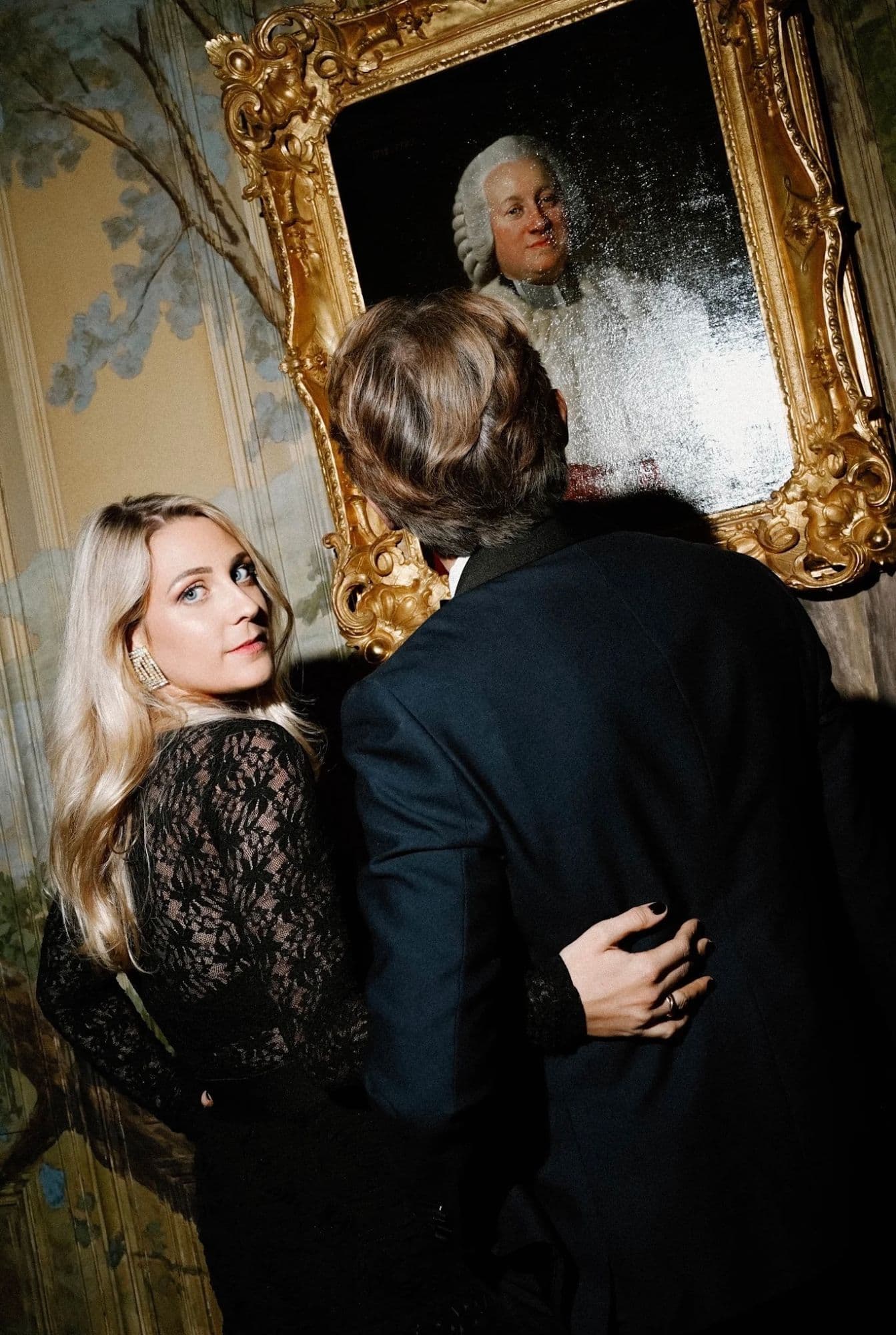 A guest in black lace beside a portrait in a gilded frame